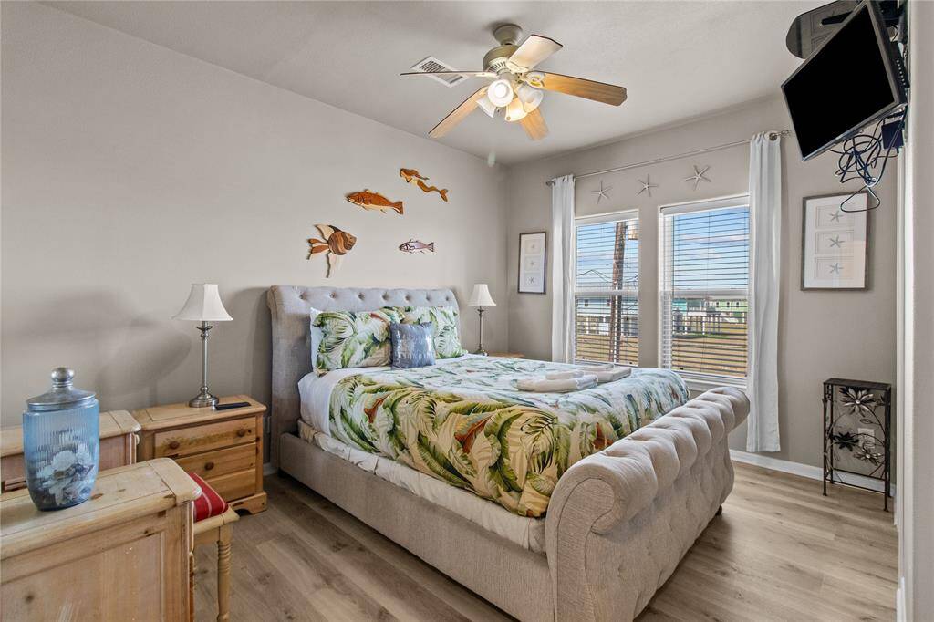 This cozy bedroom features a plush bed with tropical-themed bedding, light wood furniture, and decorative wall accents. Natural light streams through large windows, brightening the space. A ceiling fan and mounted TV add comfort and convenience.