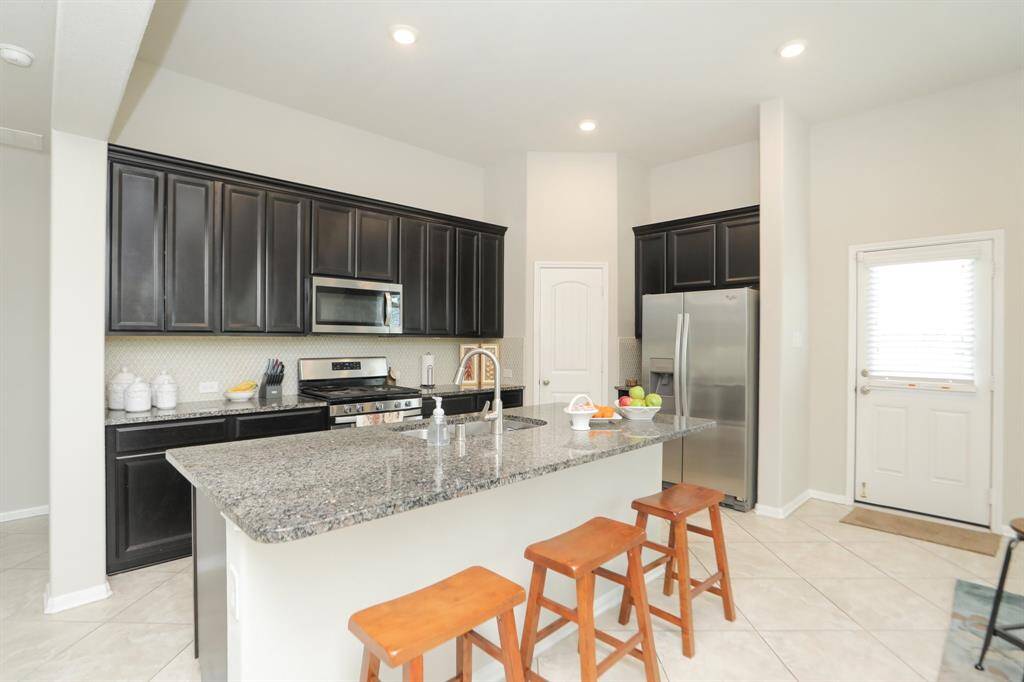This kitchen features sleek dark cabinetry, a spacious granite island with seating, and modern stainless steel appliances. The open layout is complemented by tiled flooring and ample natural light.