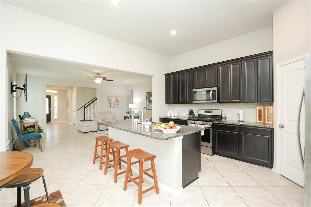 This photo showcases a bright, open-concept kitchen and living area. The kitchen features dark cabinetry, stainless steel appliances, and a large island with seating. The space flows into a cozy living room with tiled flooring and a ceiling fan, leading to a staircase and front entryway.