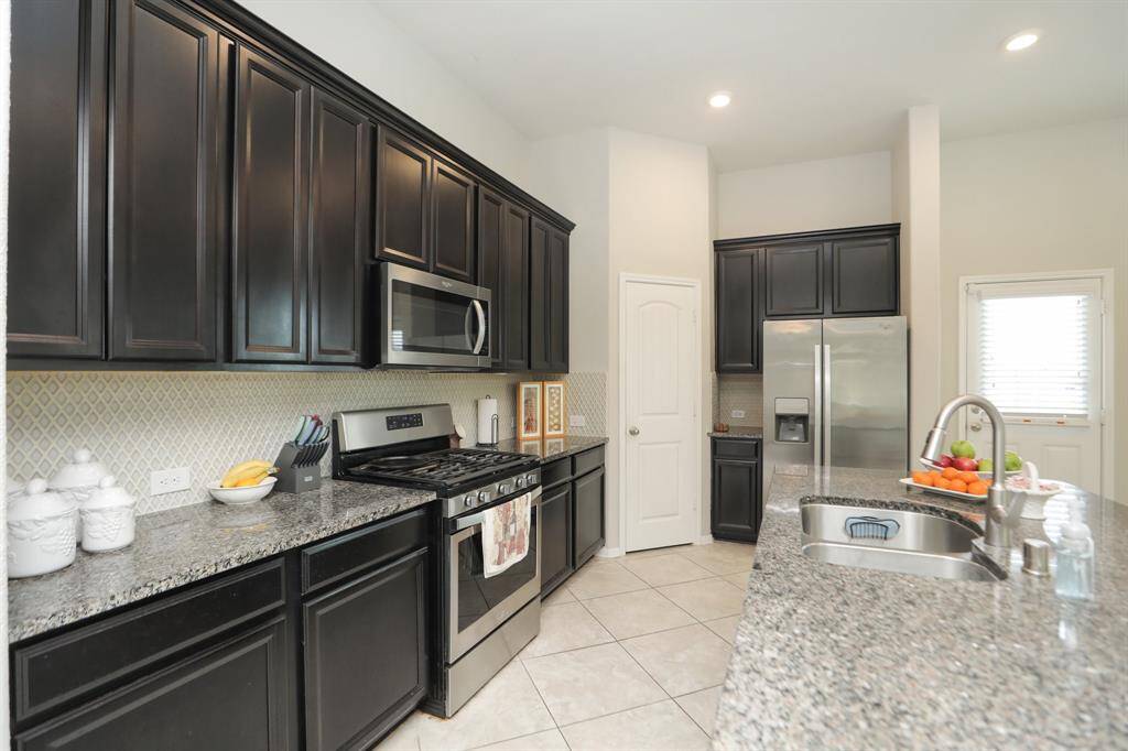 Modern kitchen with dark cabinetry, stainless steel appliances, and granite countertops. Features a gas stove, double-door refrigerator, and a spacious island with a sink. Light tile flooring and ample natural light enhance the space.