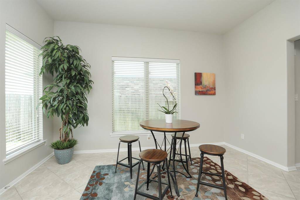 Bright dining area with a round table and four stools, featuring large windows, a potted plant, modern wall art, and a decorative rug on tiled flooring.