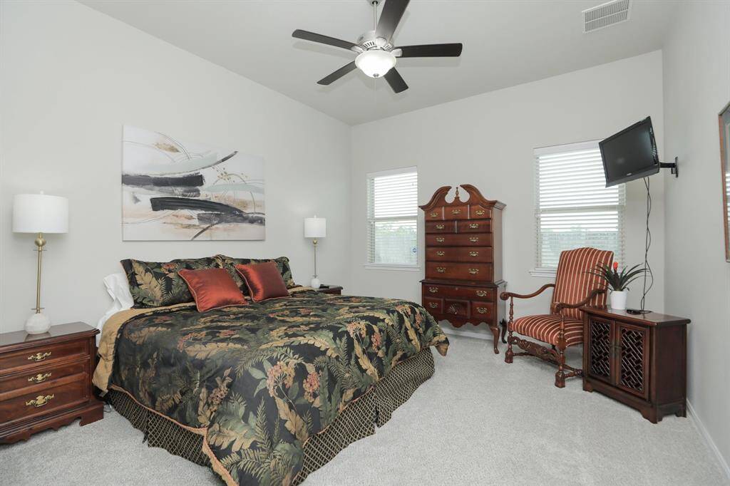 This spacious bedroom features neutral walls, a ceiling fan, and plush carpeting. It includes a large bed with elegant bedding, classic wooden furniture, and a mounted TV. Two windows provide natural light, creating a cozy, inviting atmosphere.