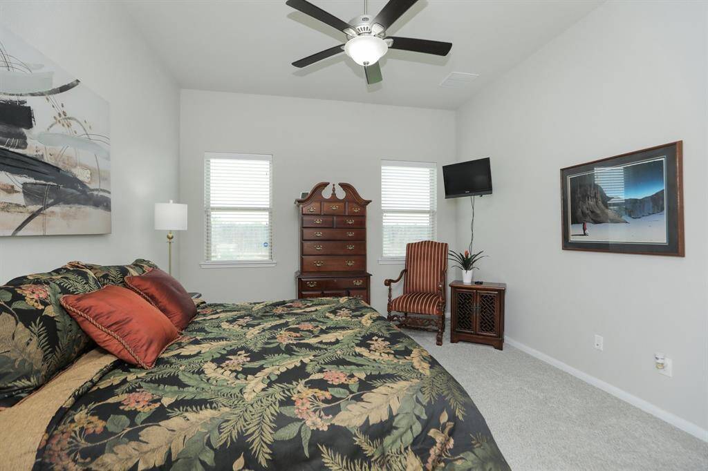This cozy bedroom features neutral walls and carpet, a ceiling fan, and natural light from two windows. It includes high ceilings and ample space for large furniture pieces.
