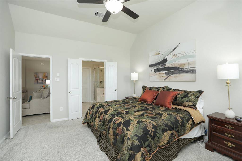 This bedroom features a spacious layout with a vaulted ceiling and neutral walls. The room connects to a bathroom with a glass shower, and there's a glimpse of a cozy living area through an open door.