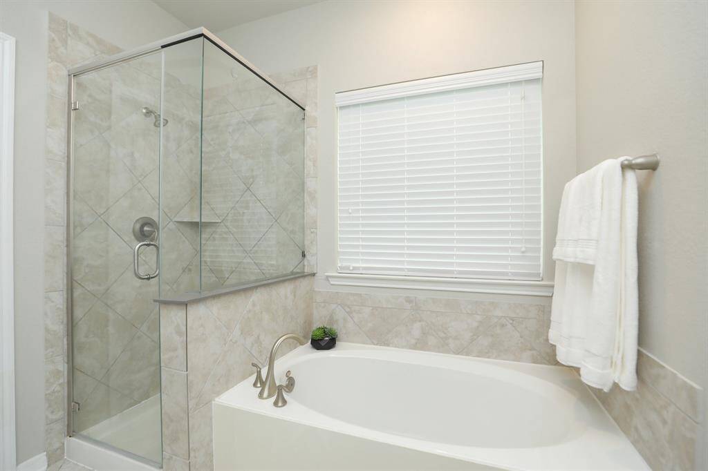 This bathroom features a modern design with a glass-enclosed shower and a spacious bathtub. Neutral tiles and a window with blinds add to the bright, clean aesthetic.
