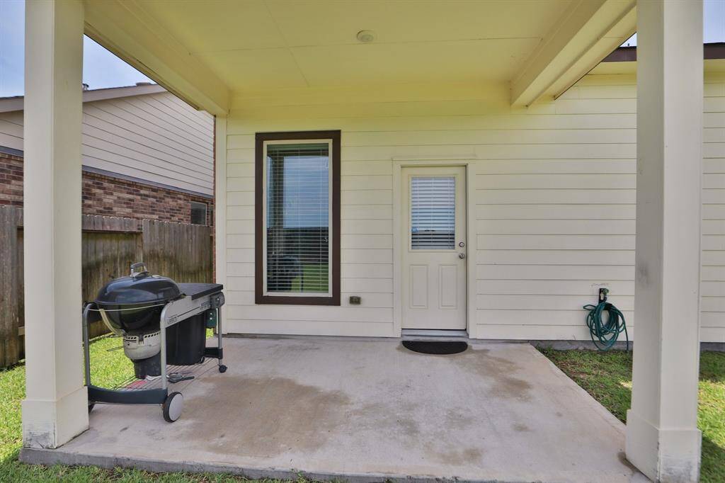 This photo shows a covered patio with a concrete floor, featuring a barbecue grill and a hose reel on the side. A door with a window and blinds leads to the interior, and there's a wooden fence providing privacy. Perfect for outdoor entertaining.
