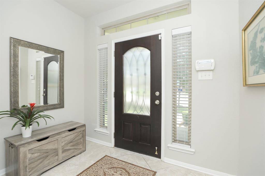 This bright entryway features a dark wood front door with frosted glass panels, flanked by tall windows with blinds. The space is accented by a large mirror, a decorative bench with storage, and light-colored tile flooring, creating an inviting and functional entrance.