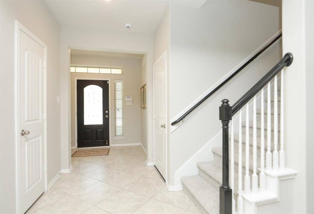 This  shows a bright, welcoming entryway with tiled floors, a dark front door with glass panels, and a staircase with a black handrail and white balusters leading upstairs. The space is painted in a light, neutral color, enhancing the open feel.
