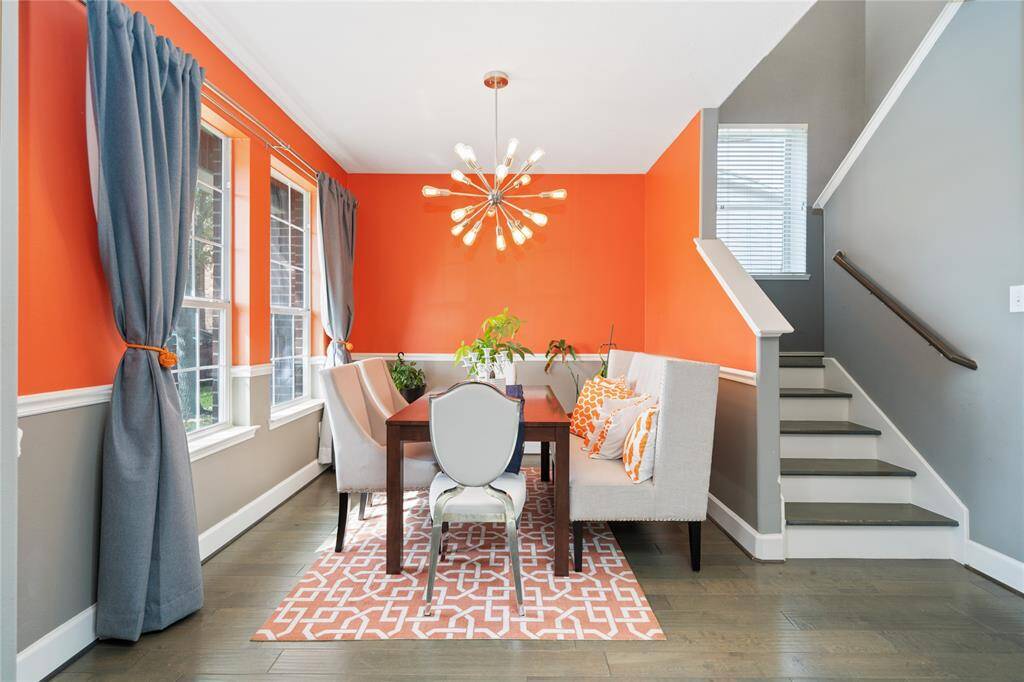 This vibrant dining area features bright orange walls and stylish gray accents, complemented by large windows with gray curtains. It includes a modern light fixture, a cozy dining setup with a cushioned bench, and an accent chair. The space is adjacent to a staircase, offering a contemporary and inviting atmosphere.