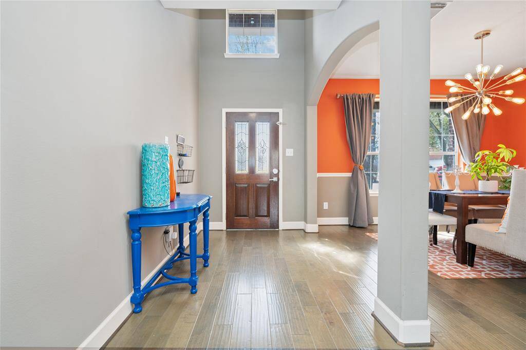 Bright and inviting entryway with high ceilings and hardwood floors. Features a striking blue console table and leads into a dining area with orange accent walls and modern lighting. Natural light fills the space through large windows.