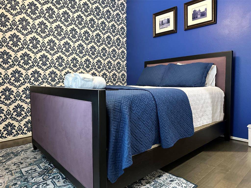 This bedroom features a stylish design with a blue accent wall and a patterned wallpaper. It includes a modern bed with deep blue bedding, complemented by framed artwork. The wood flooring and a decorative rug add warmth and character.