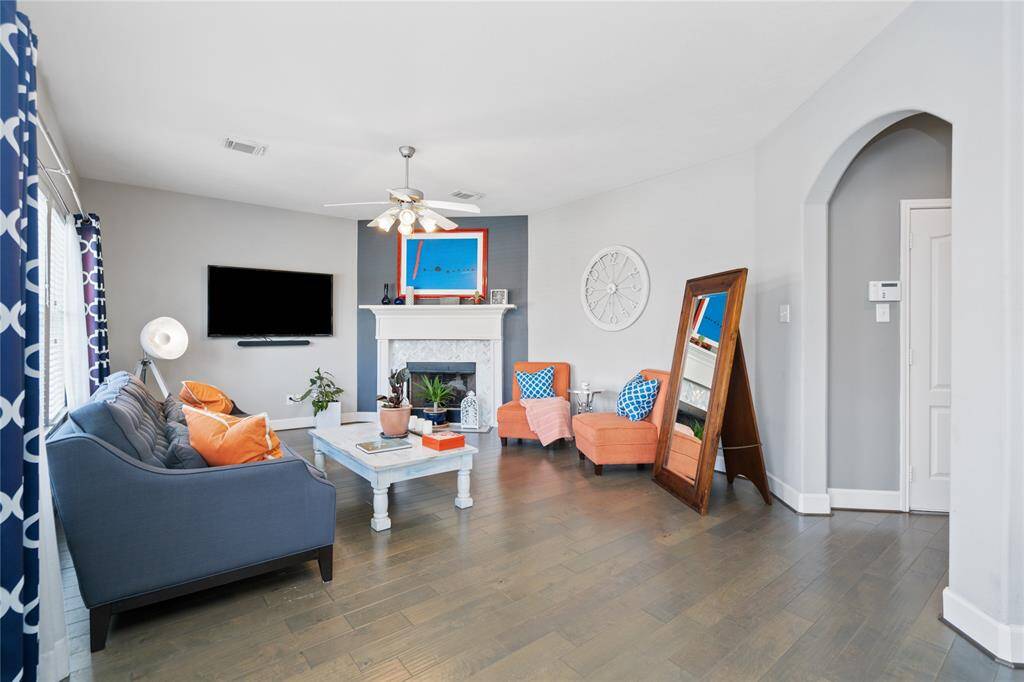 Bright and inviting living room featuring hardwood floors, a cozy fireplace, and a modern ceiling fan. The space is furnished with a comfortable sofa and vibrant chairs, complemented by stylish decor and plentiful natural light.