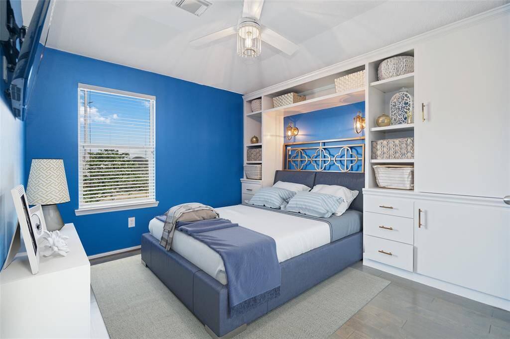 This cozy bedroom features a modern blue accent wall, built-in shelving, and a comfortable bed. Ample natural light flows through the window, and the room includes stylish overhead lighting and a ceiling fan.