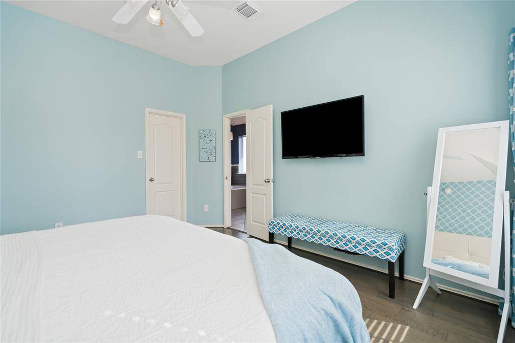 Bright bedroom with light blue walls, featuring a ceiling fan, wall-mounted TV, and a patterned bench. It includes a full-length mirror and an en-suite bathroom.