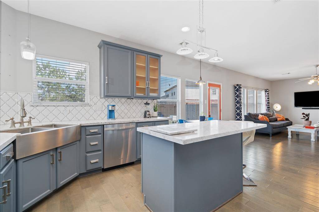 This photo showcases a bright, modern kitchen and living area. The kitchen features sleek blue cabinets, a stylish backsplash, stainless steel appliances, and a large island with pendant lighting. The open layout flows into a cozy living space with ample natural light and contemporary decor.
