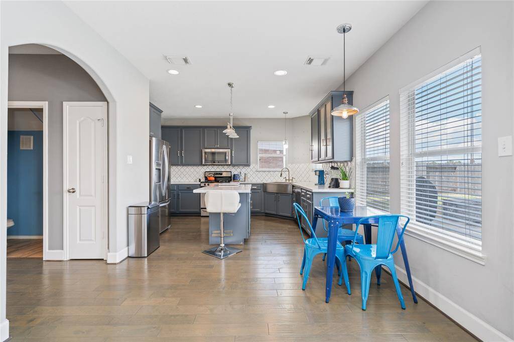 This photo showcases a modern kitchen with gray cabinetry, stainless steel appliances, and a central island with bar seating. It features a cozy breakfast nook with bright blue chairs next to large windows, allowing plenty of natural light. The space has an open and inviting layout, ideal for family gatherings.