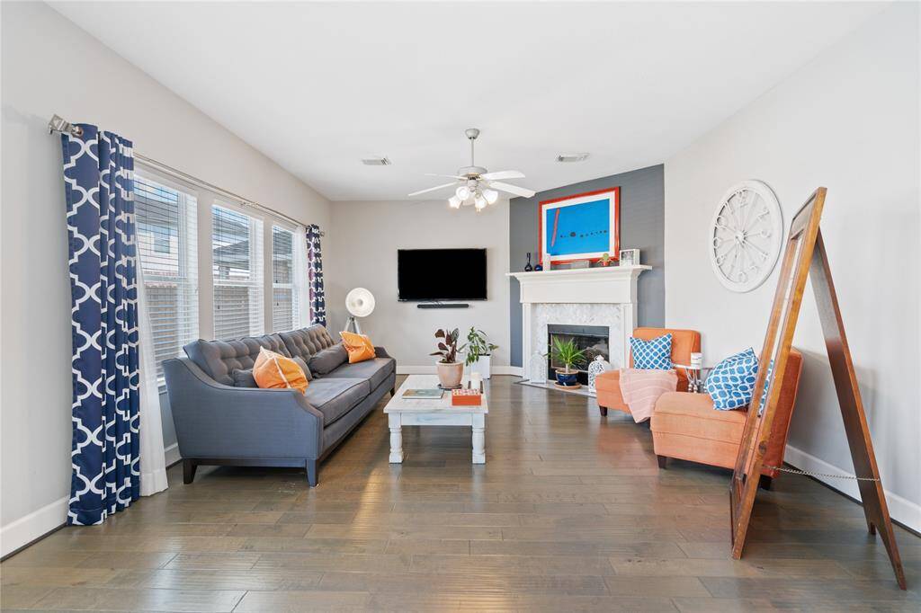 Bright and inviting living room featuring hardwood floors, a cozy fireplace, and stylish decor. The space includes a comfortable sofa, modern accent chairs, and large windows with patterned curtains, allowing plenty of natural light. Perfect for relaxation and entertaining.