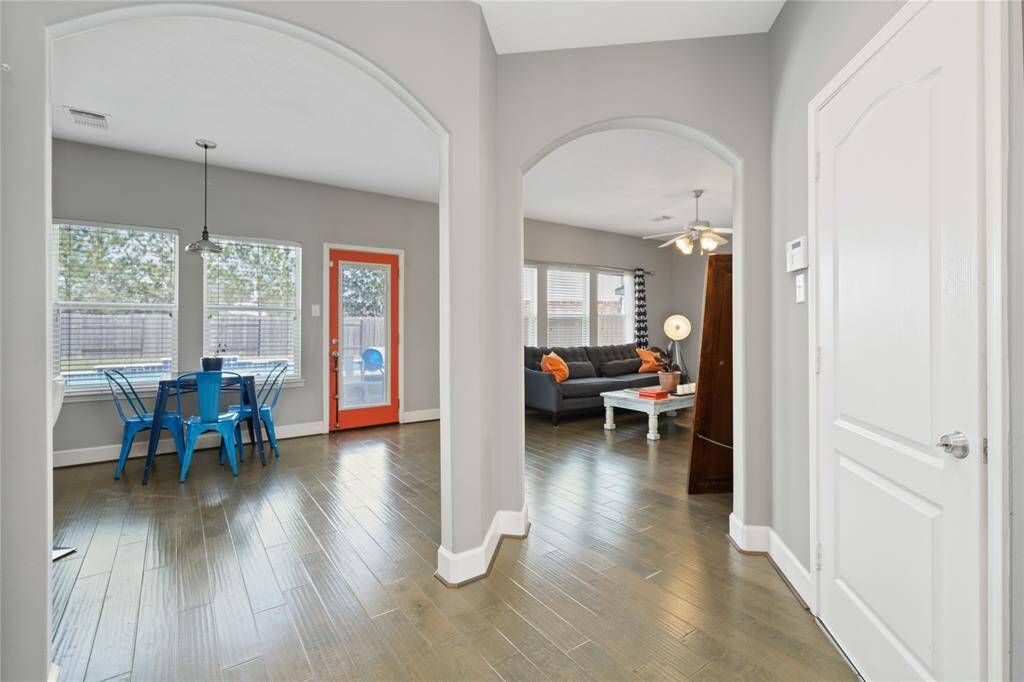 Bright and inviting open-concept space with hardwood floors, featuring a cozy living area with a ceiling fan and a dining area with vibrant blue chairs. Large windows provide plenty of natural light, complementing the modern gray walls and offering a view of the backyard.