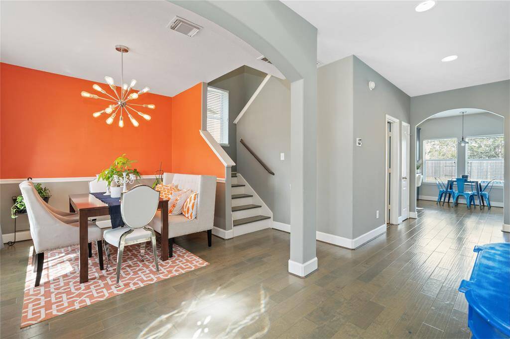 This photo showcases a modern dining area with a vibrant orange accent wall and stylish light fixture. It features a cozy dining set with a bench, adjacent to a staircase with neutral gray walls. The space leads to another room with large windows and ample natural light, creating a warm and inviting atmosphere.