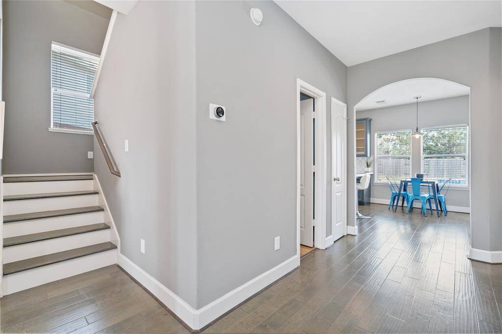This photo showcases a bright, modern interior with hardwood floors and neutral walls. It features a staircase, a hallway leading to a kitchen area with large windows, and a cozy dining space with blue chairs.
