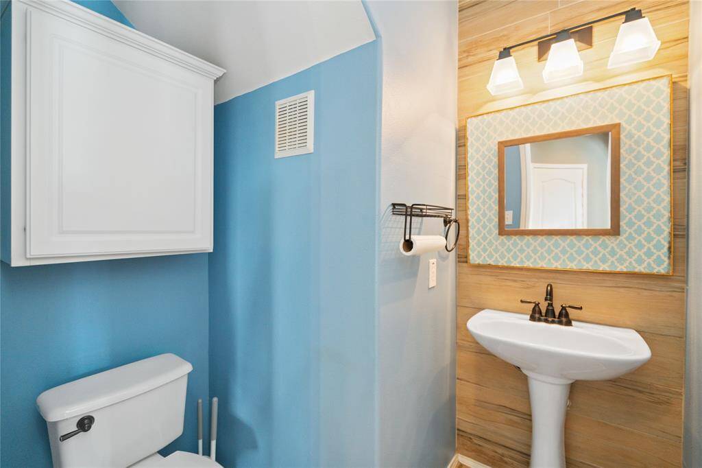This bathroom features a cozy design with light blue walls, a pedestal sink, and a toilet. It includes a decorative mirror with a patterned frame, modern lighting, and a storage cabinet for convenience.