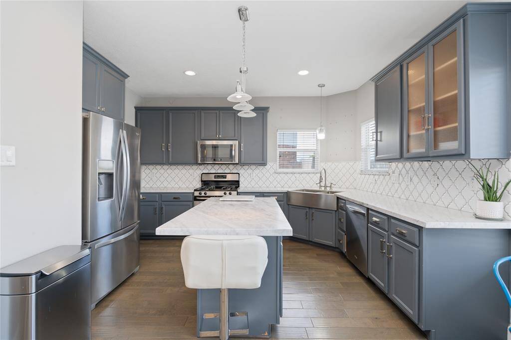 Modern kitchen with sleek gray cabinets, stainless steel appliances, and a central island with a marble countertop. The room features a farmhouse sink, stylish pendant lighting, and a geometric tile backsplash, creating a bright and inviting space.