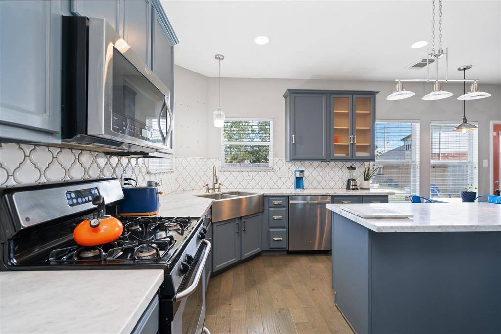 Modern kitchen with sleek gray cabinets, stainless steel appliances, and a spacious island. Features a stylish backsplash, pendant lighting, and ample natural light from large windows. Perfect for cooking and entertaining.