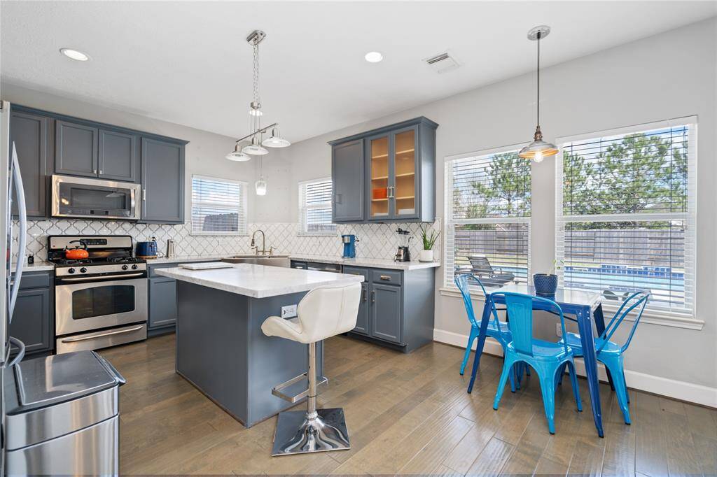 Bright and modern kitchen with sleek dark cabinets, stainless steel appliances, and a central island. The dining area features a chic table with blue chairs, and large windows provide natural light and a view of the backyard with a pool.