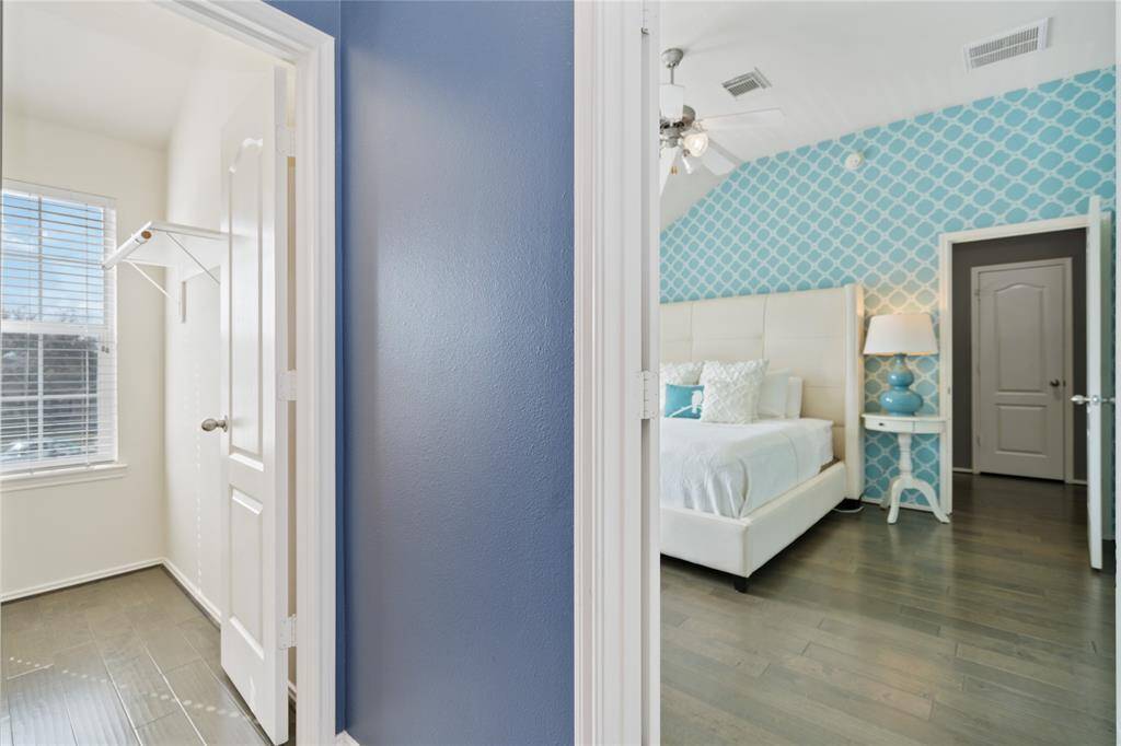 This photo shows a bright, stylish bedroom with a modern blue accent wall and hardwood flooring. It features a cozy bed with decorative pillows and a small bedside table with a lamp. On the left, there's a walk-in closet with a window, providing natural light.