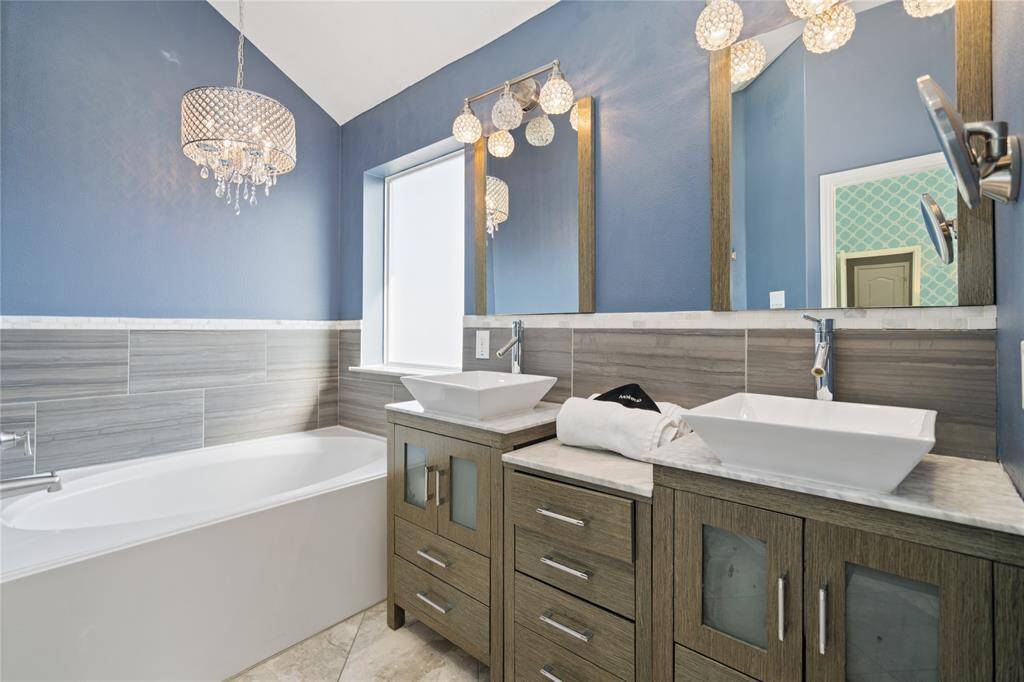 This modern bathroom features a stylish blue accent wall, elegant chandelier lighting, dual vessel sinks with a wood vanity, and a spacious bathtub with sleek gray tile surround.
