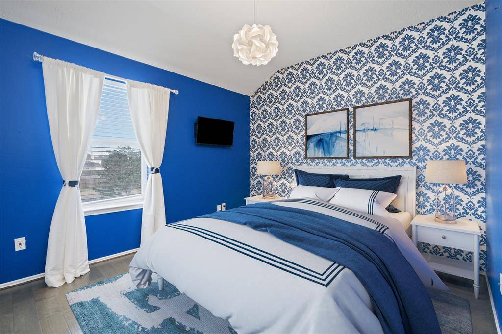 This bedroom features a bold blue and white color scheme with a patterned accent wall. It includes a cozy bed with matching linens, bedside tables with lamps, a wall-mounted TV, and a window with white curtains, creating a bright and inviting space.