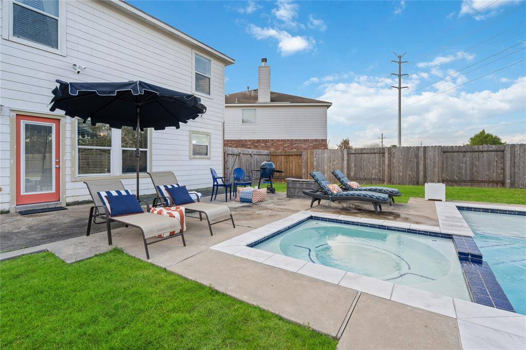 This backyard features a spacious patio with comfortable lounge chairs, a large umbrella for shade, and a sparkling pool with a hot tub, perfect for relaxation and entertaining. The area is enclosed by a privacy fence, offering a serene outdoor retreat.
