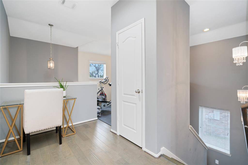 This image showcases a bright, modern upstairs hallway with wooden flooring and a neutral color palette. It features a small desk area under a hanging light fixture, adjacent to a room with exercise equipment. The staircase is visible with elegant lighting and a window providing natural light.