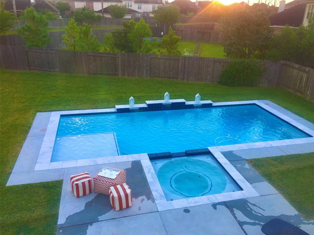 This backyard features a modern rectangular pool with a connected circular hot tub, surrounded by a spacious patio. The area includes a grassy lawn and is enclosed by a wooden fence, offering privacy and a serene atmosphere.
