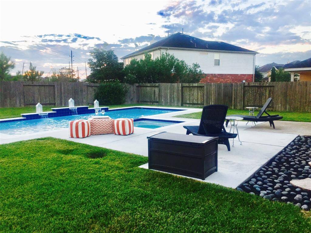 This photo showcases a spacious backyard featuring a well-maintained pool with water features. It includes a surrounding patio area with lounge chairs and a seating area, offering a perfect space for relaxation and outdoor entertaining. The yard is enclosed by a wooden fence, providing privacy.