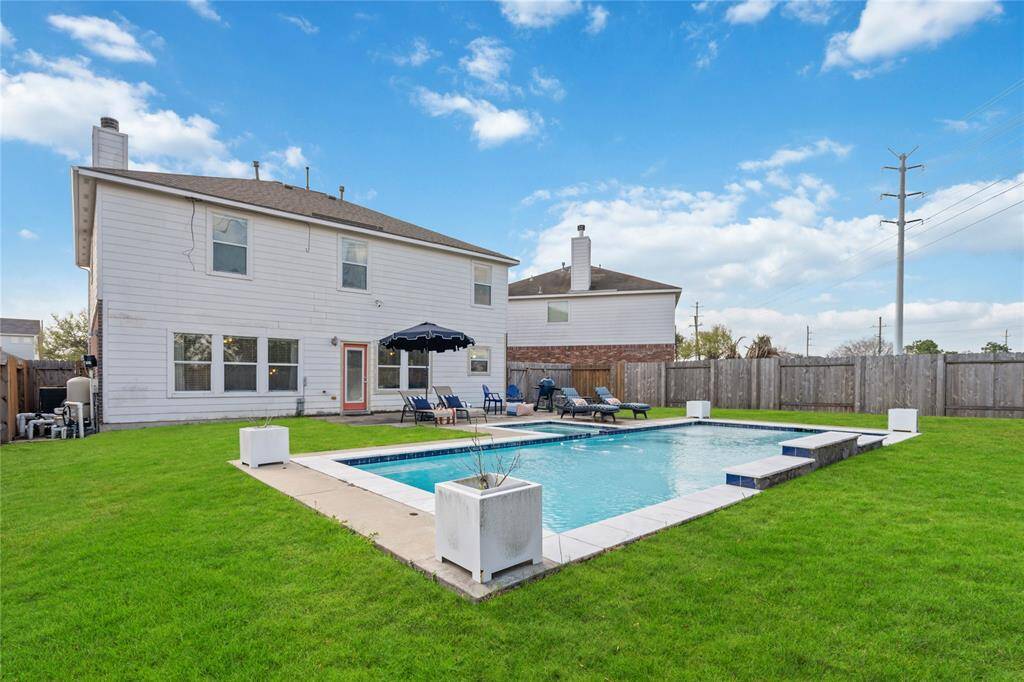 This photo showcases a spacious backyard featuring a large, inviting swimming pool surrounded by well-maintained green grass. The area includes lounge chairs for relaxation and is bordered by a wooden fence, offering privacy. The house exterior is visible, with multiple windows and a clean, white finish.