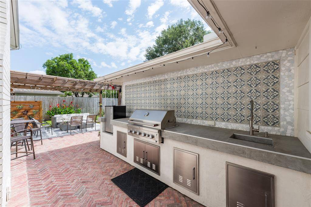 A gorgeous outdoor kitchen with a built-in grill, sink, and patterned tile backsplash. The area features a pergola-covered patio with seating, perfect for entertaining, and a neat brick floor. Lush greenery and a privacy fence complete the inviting backyard space.