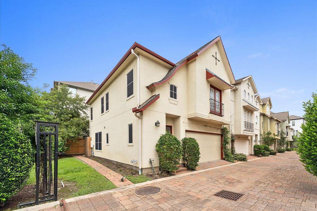 This photo showcases a charming, multi-story townhouse with a stucco exterior and a gable roof. It features a neatly paved driveway, a two-car garage, and a small, well-maintained side yard with greenery. The home is part of a row of similar houses, indicating a cohesive neighborhood setting.
