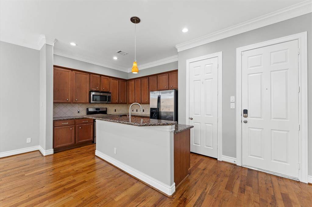 This kitchen features hardwood floors, sleek granite countertops, and modern stainless steel appliances. It has warm wooden cabinets, a central island with a sink, and recessed lighting, creating a bright and inviting space.