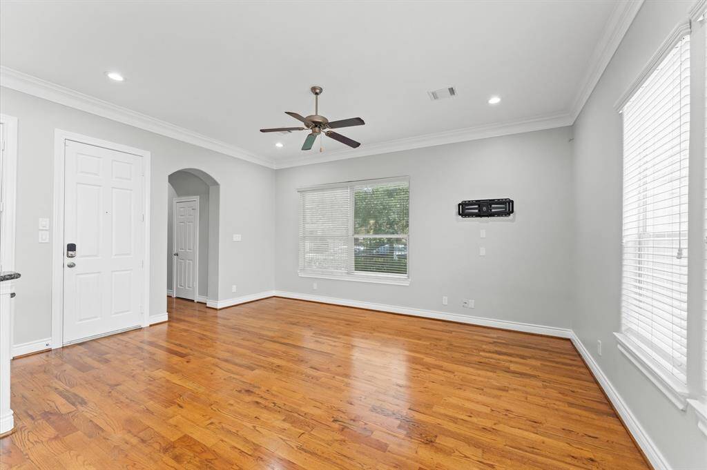 This spacious living area features polished hardwood floors, neutral gray walls, and ample natural light from large windows. The room includes recessed lighting, a ceiling fan, and an archway leading to an adjacent space, offering a modern and inviting atmosphere.