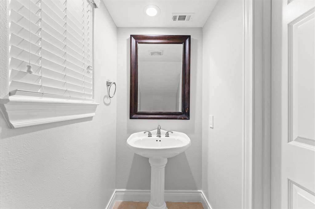 This is a compact, well-lit bathroom with a pedestal sink, a dark-framed mirror, and a window with blinds. The space is clean and bright, ideal for a guest or powder room.
