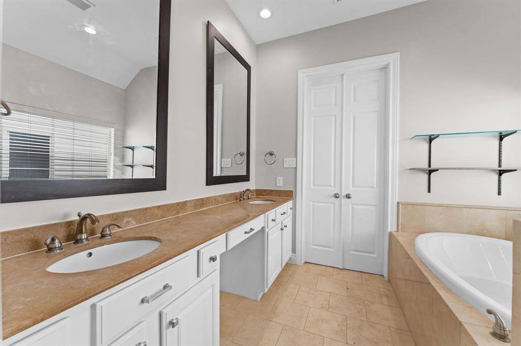 This bathroom features a double vanity with elegant brown countertops and modern fixtures, a large mirror, and ample storage in white cabinets. It has a luxurious soaking tub, a neutral color palette, and natural light from a window with blinds.