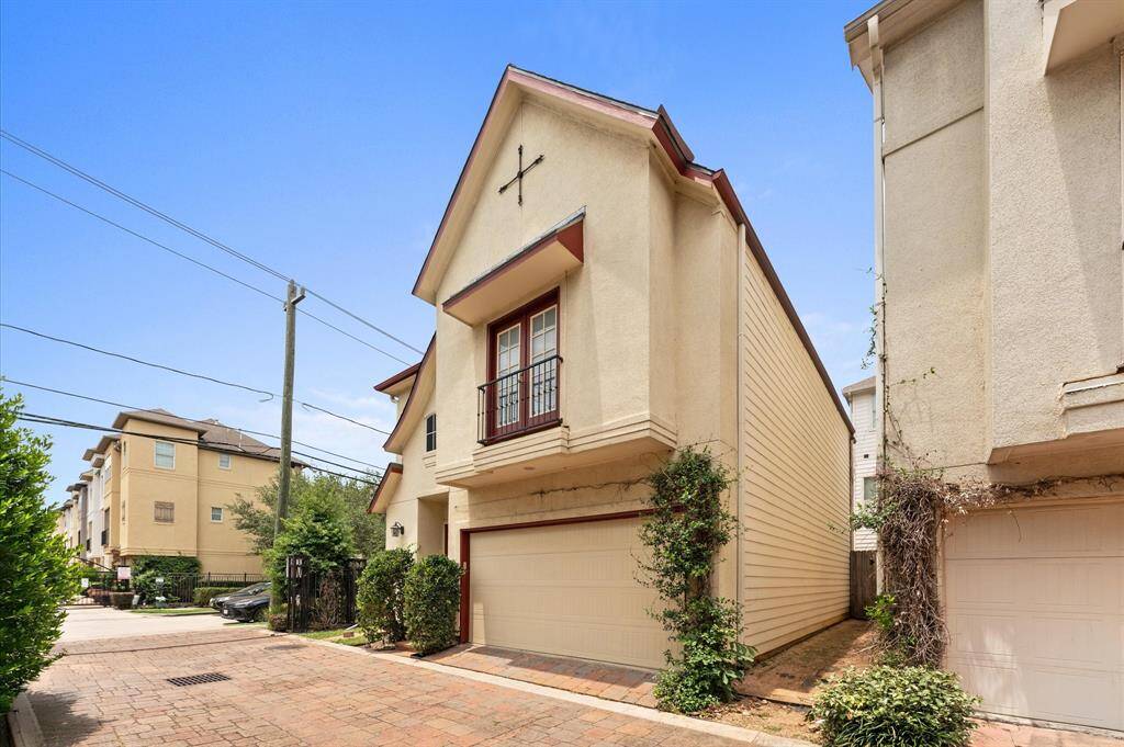 This photo shows a modern, multi-story townhouse with a two-car garage and a small balcony. The exterior features light-colored stucco with red trim, situated in a well-maintained, paved residential area.