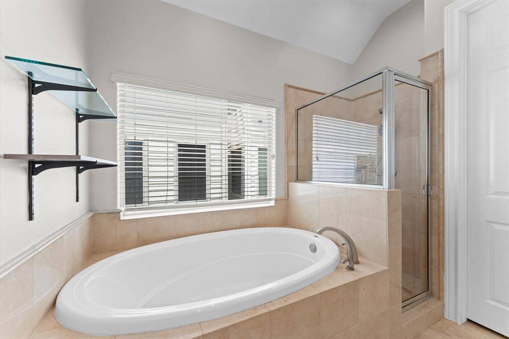 This bathroom features a spacious oval bathtub and a separate glass-enclosed shower. It has neutral tiled walls and floors, a large window with blinds, and modern shelving for storage. The overall design is bright and airy.