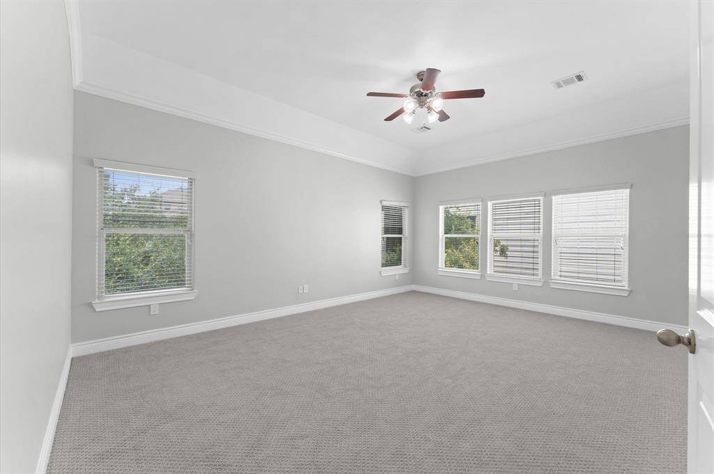 Spacious, light-filled primary bedroom with neutral walls and carpet, featuring multiple windows with blinds, a ceiling fan, and crown molding.