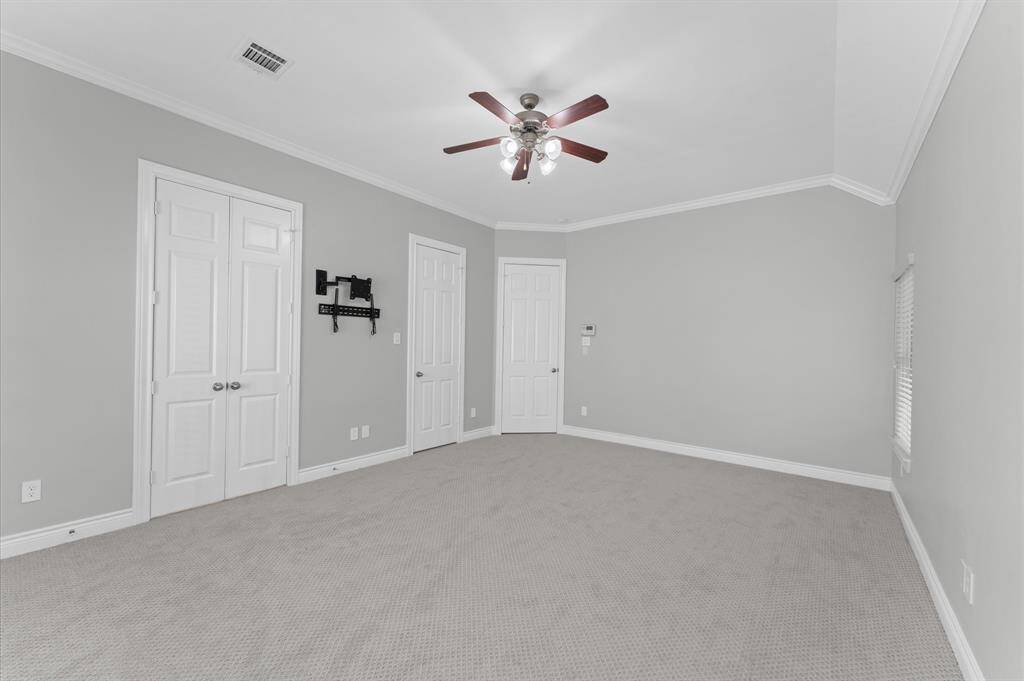This is a spacious, carpeted room with neutral gray walls, crown molding, and a ceiling fan. It features two white doors and a wall bracket for a TV. Perfect as a bedroom or office space.