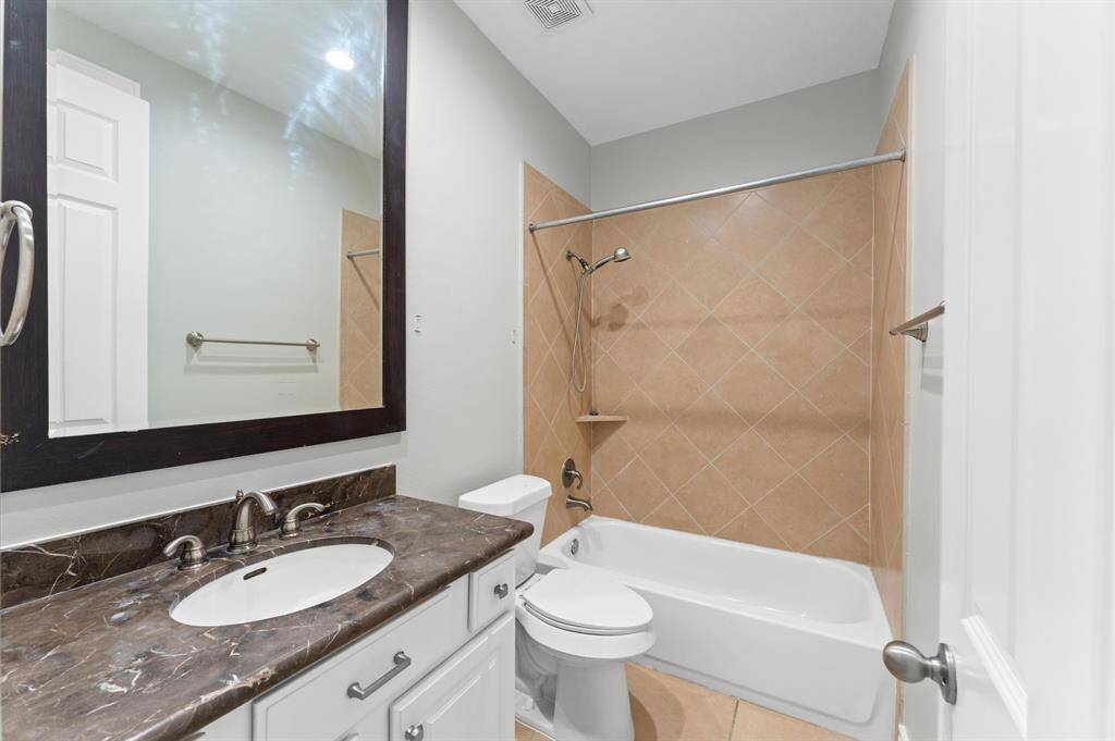 This bathroom features a sleek design with a granite countertop, a large framed mirror, and a bathtub with beige tile surround. It includes modern fixtures and a neutral color palette, providing a clean and inviting space.