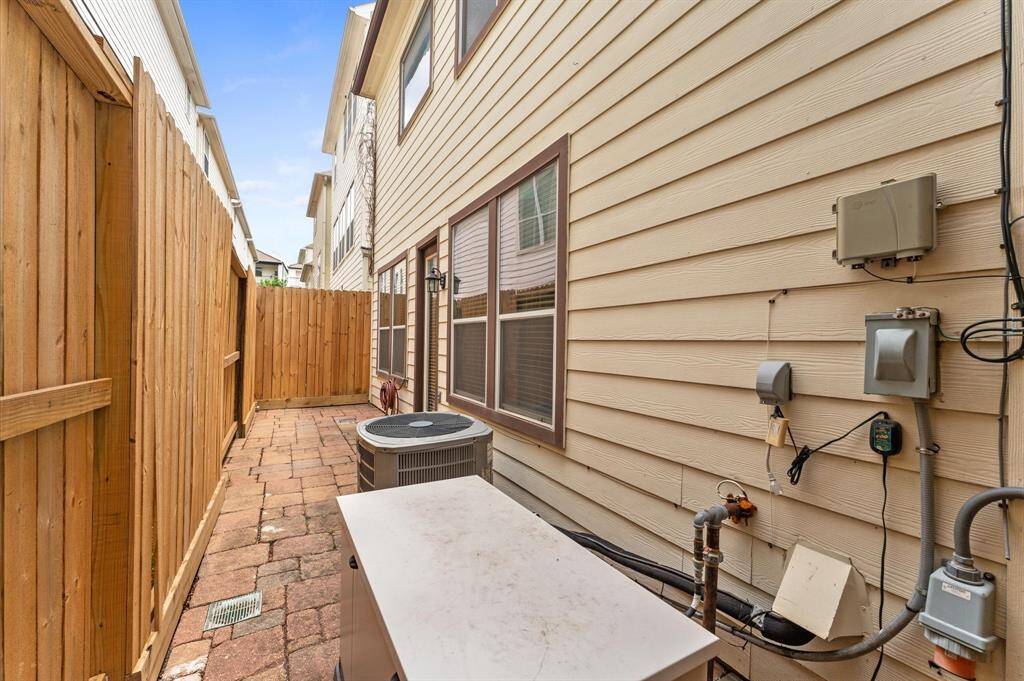This photo shows a narrow side yard with a paved walkway, bordered by a wooden fence and the exterior wall of a house. It features an air conditioning unit and utility connections, providing essential outdoor space for maintenance access.