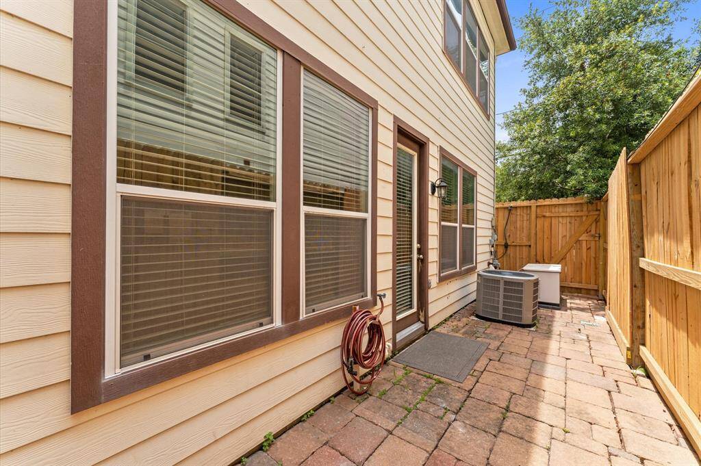 This photo shows a narrow side yard with a paved walkway, bordered by a wooden fence. The house exterior features large windows and a door with blinds. An air conditioning unit is visible, along with a garden hose.