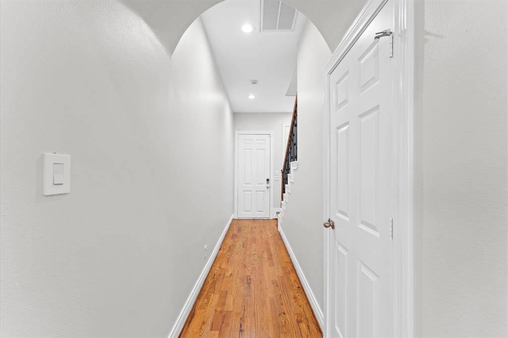 This photo shows a bright, welcoming hallway with polished hardwood floors, neutral-toned walls, and recessed lighting. The hallway features an arched ceiling and leads to a staircase with a wooden railing. There are white doors on the right side, adding to the clean and modern look.